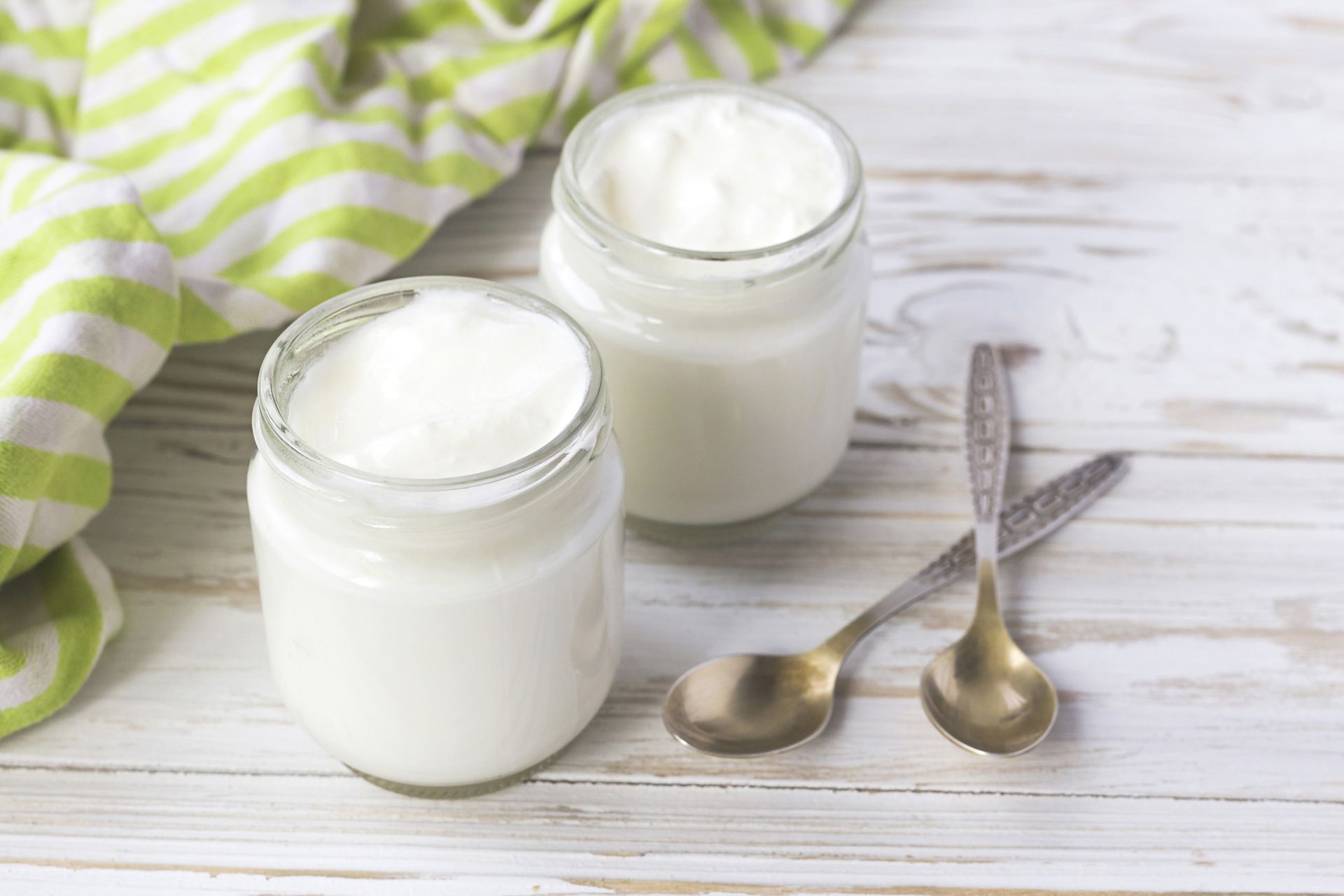 Homemade yogurt in glass jar with spoons and green dishcloth on wooden table.