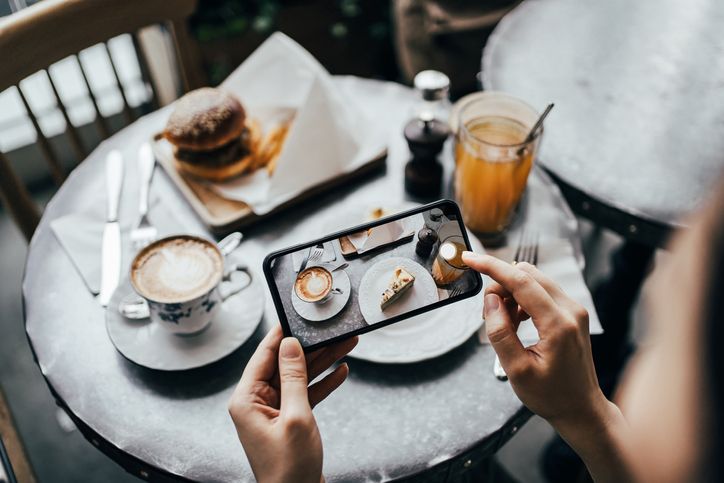 Overhead view of young woman taking photo of freshly served food, burger with fries and cream cake with smartphone and sharing to social media before eating in a stylish coffee shop
