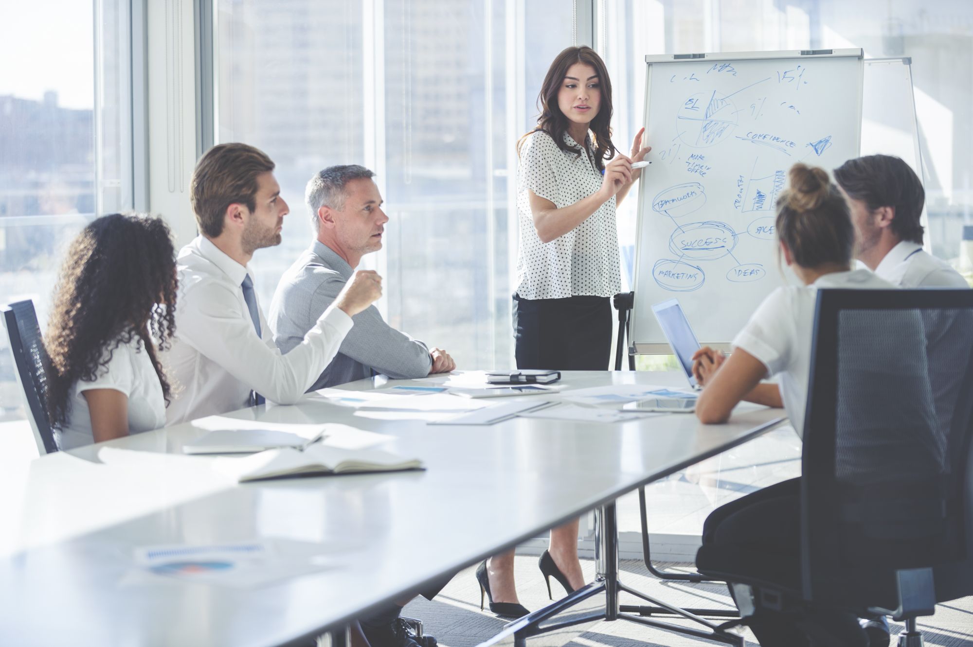 Woman giving a presentation to her team. She is using a whiteboard with charts and graphs. She is talking. They are in an office boardroom at the table with laptop computers and paperwork. She is smiling. Men and women in the group.